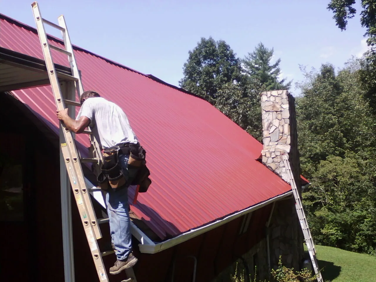 Roofer on a ladder beside a red metal roof and stone chimney — Soffit & Fascia Repair by Roofing Brothers in New Laguna